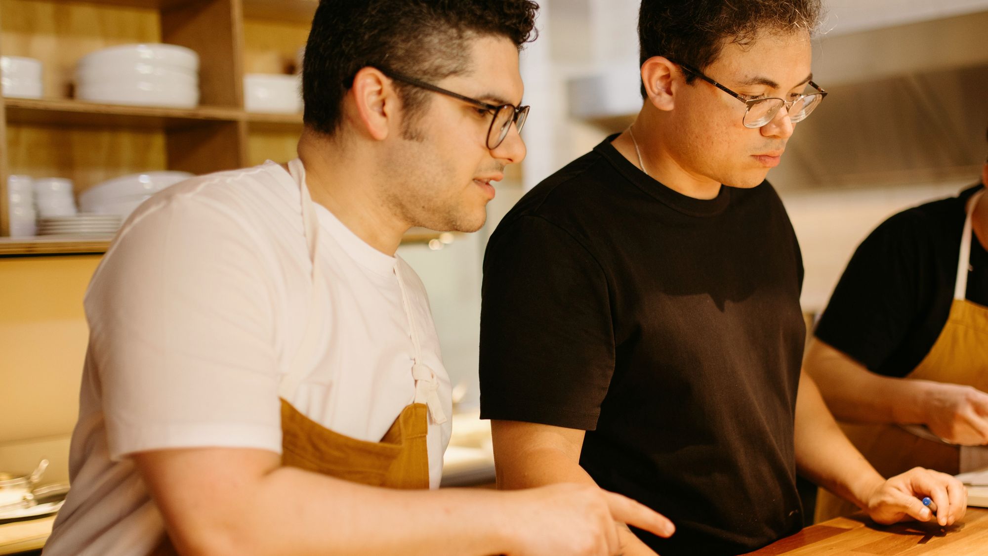 Un équipier explique un geste à un collègue au plan de travail d’une cuisine, pendant la préparation