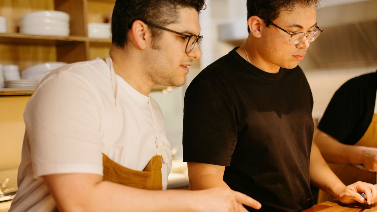 Un équipier explique un geste à un collègue au plan de travail d’une cuisine, pendant la préparation