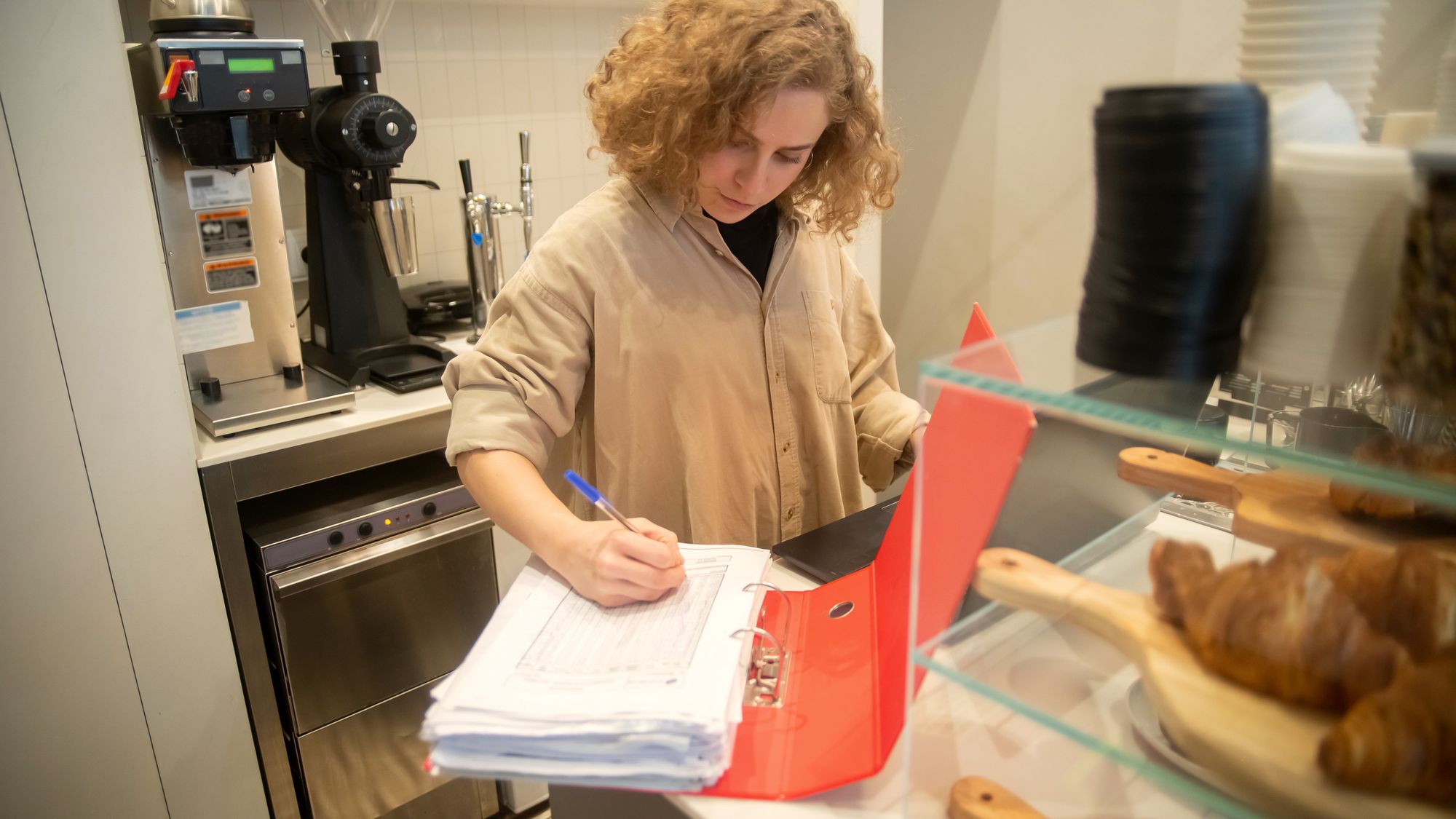 Une gérante remplit un classeur de chiffres au comptoir de son établissement, à côté des viennoiseries