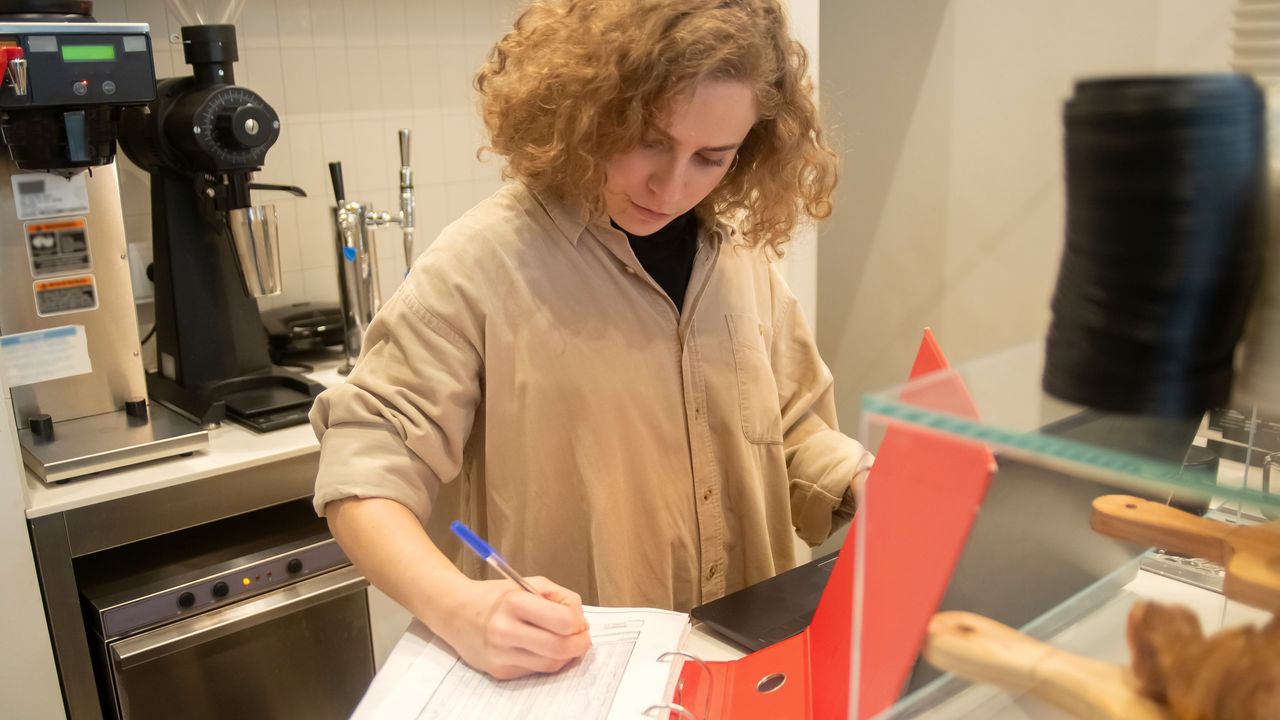 Une gérante remplit un classeur de chiffres au comptoir de son établissement, à côté des viennoiseries