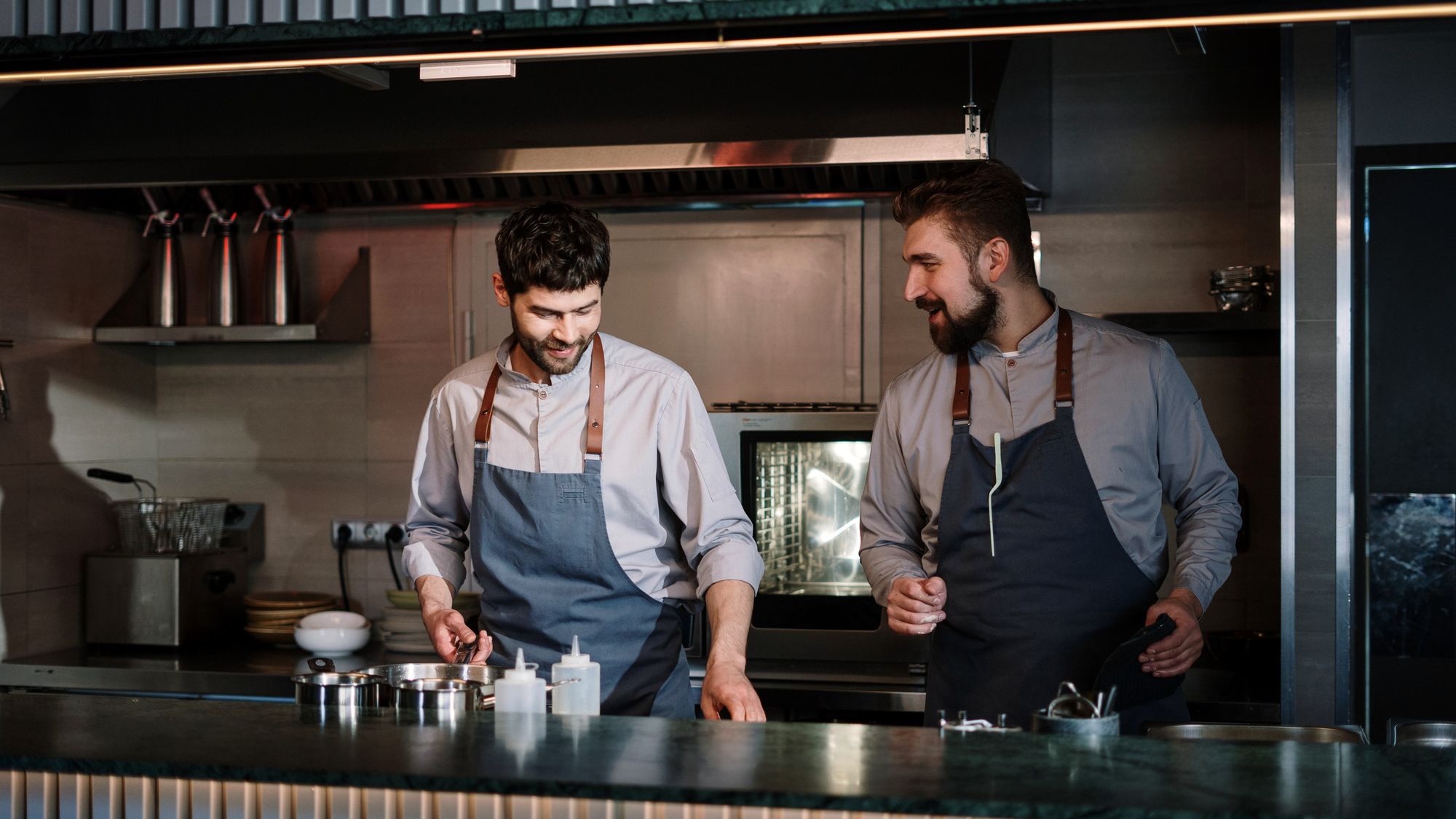 Deux équipiers en tablier échangent à leur poste dans une cuisine ouverte, pendant la mise en place