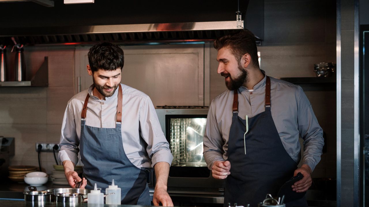Deux équipiers en tablier échangent à leur poste dans une cuisine ouverte, pendant la mise en place