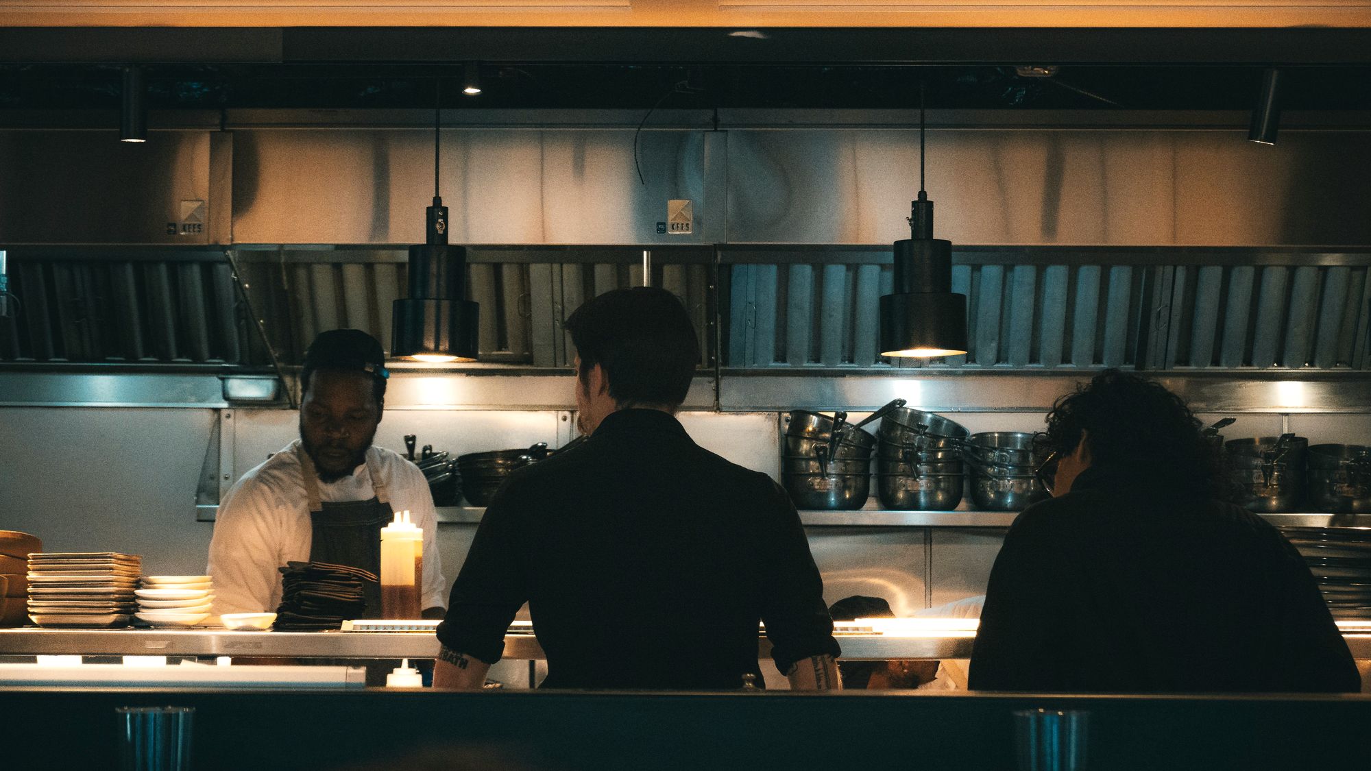 Équipe de cuisine au passe pendant le service du soir, sous les lampes, assiettes en préparation