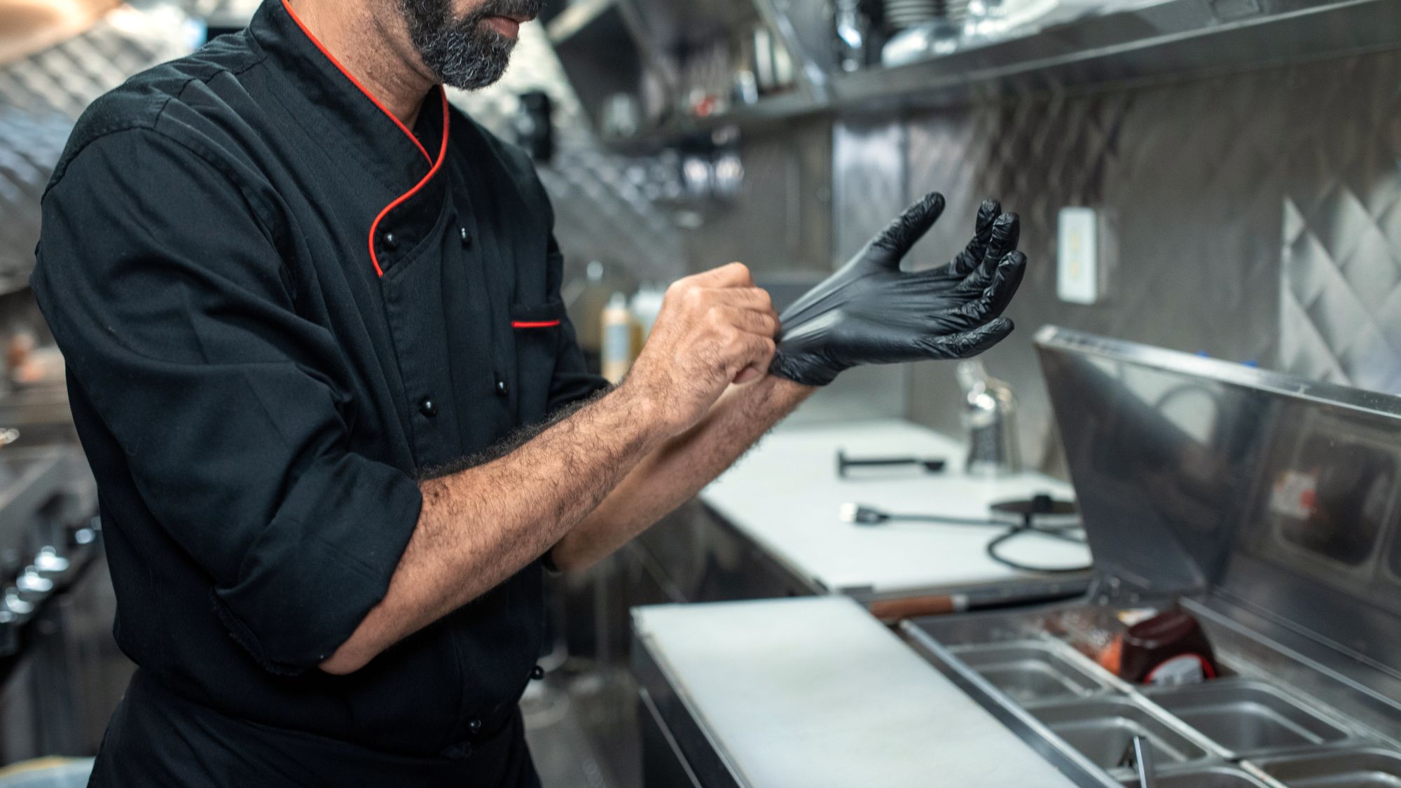 Cuisinier qui enfile des gants noirs à son poste, devant les bacs inox de la saladette réfrigérée