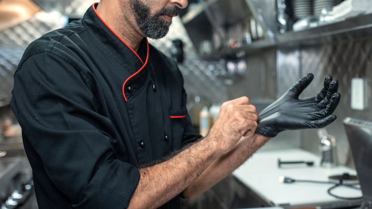 Cuisinier qui enfile des gants noirs à son poste, devant les bacs inox de la saladette réfrigérée